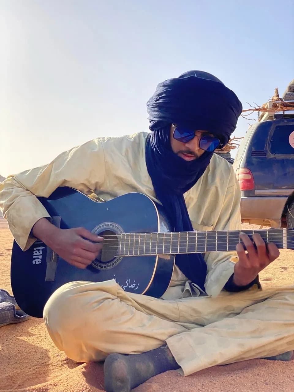 A man in a desert veil playing guitar on the sand beside a 4×4
