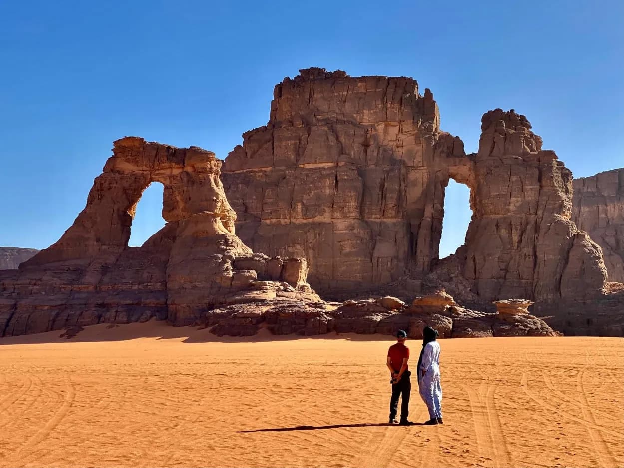 A traveller and a veiled guide beneath a sandstone arch in the Algerian Sahara